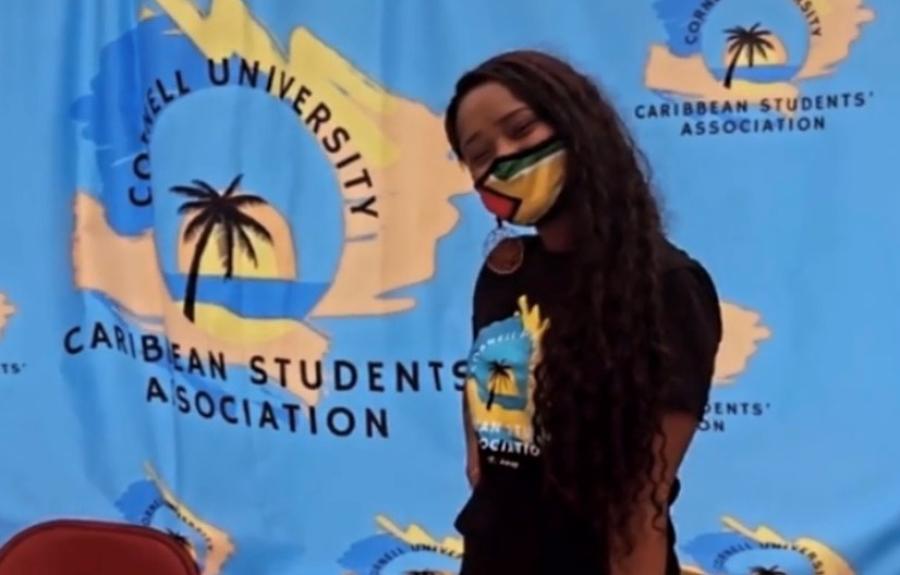 A college student in a mask in front of a Carribean Students' Association banner.